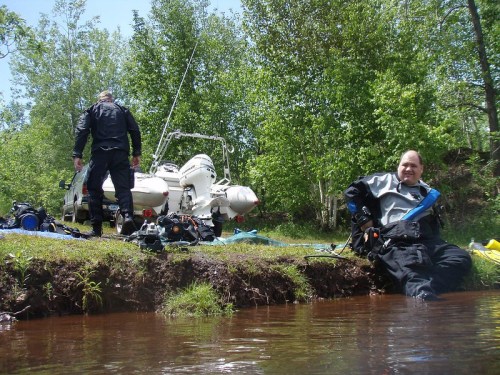 Two divers prepare for an outing by a river, with a boat and gear visible in a lush, green setting.