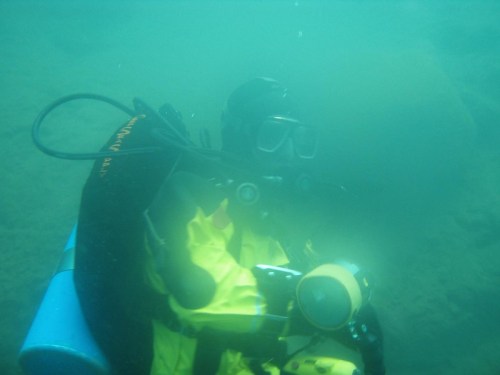 A diver in a yellow and black wetsuit underwater, holding a camera, surrounded by murky water.