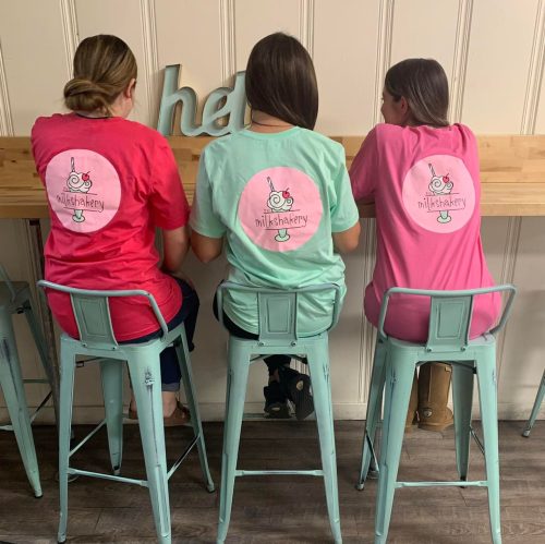 Three people sit at a counter wearing colorful t-shirts with a milkshake logo, facing away from the camera.