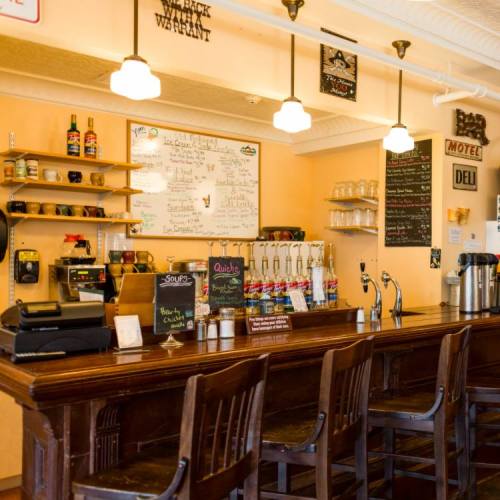 A cozy bar with wooden stools, a menu board, and shelves displaying drinks and glassware.