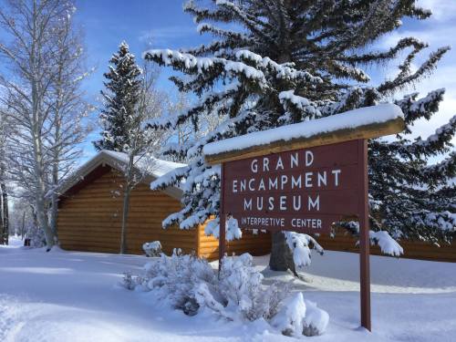 Sign for Grand Encampment Museum and Interpretive Center surrounded by snow-covered trees and a winter landscape.
