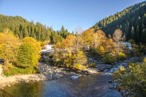A serene river flows through a forested area with autumn foliage and a small building nestled among the trees.