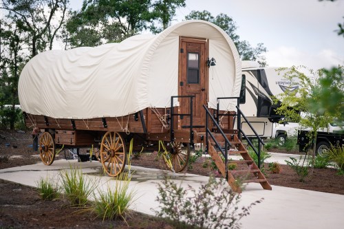 A wooden covered wagon on wheels, featuring a door and steps, set in a landscaped area with greenery.