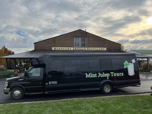 A black tour bus with "Mint Julep Tours" logo parked in front of the Kentucky Artisan Distillery building.