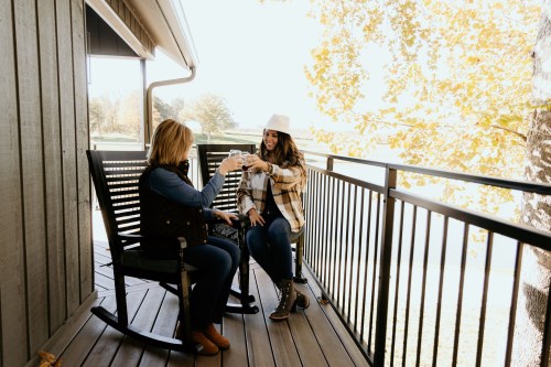Two women sit on a balcony, enjoying drinks and chatting, surrounded by trees and a sunny outdoor view.