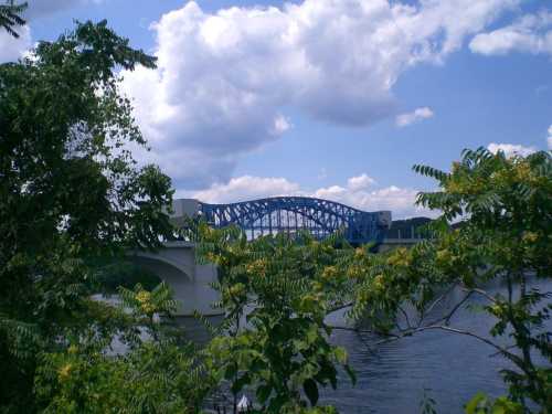 A blue bridge spans a river, framed by green trees and a partly cloudy sky.