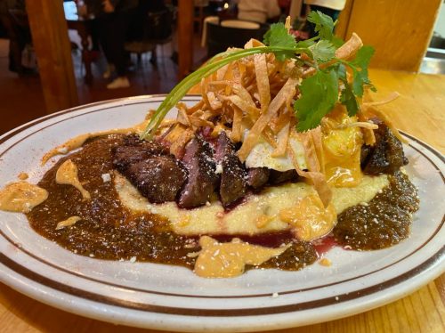 A plate of steak on creamy grits, topped with fried onions, cilantro, and a drizzle of sauce.