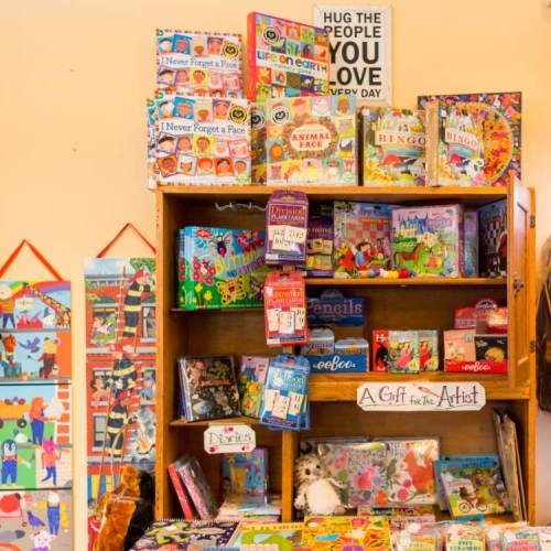 A wooden shelf filled with colorful games, puzzles, and art supplies, with a sign reading "A Gift for The Artist."