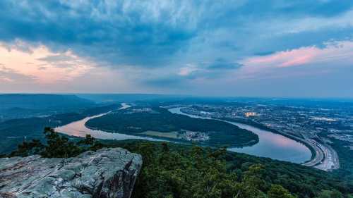 A panoramic view of a river winding through green hills and a city, under a cloudy sky at dusk.