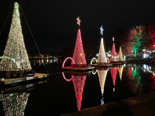 Colorful illuminated Christmas trees reflected in a calm lake at night, surrounded by festive decorations.