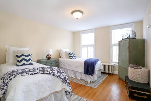Bright bedroom with two twin beds, decorative pillows, a green wardrobe, and large windows letting in natural light.