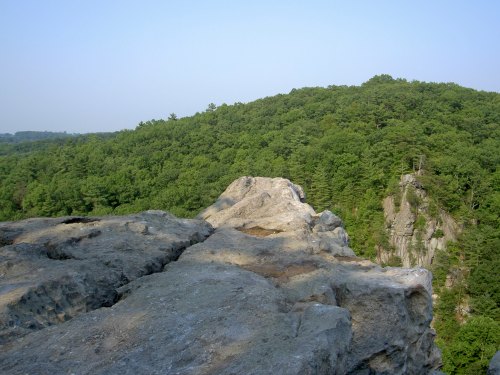 A rocky outcrop overlooks a lush green forested hillside under a clear blue sky.
