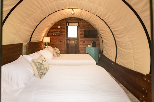 Cozy interior of a glamping tent featuring two beds, wooden accents, and a small TV. Natural light from a window.