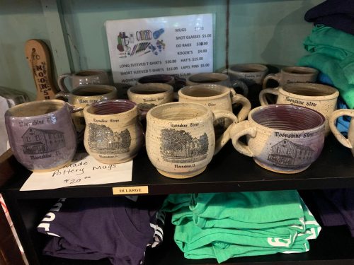 A display of handmade pottery mugs featuring scenic designs, priced at $20 each, on a shelf with T-shirts.