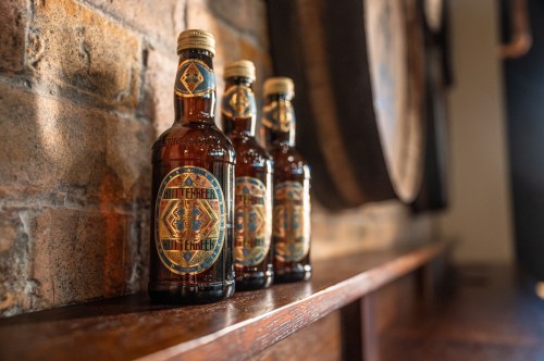 Three decorative bottles of beer on a wooden shelf against a brick wall.