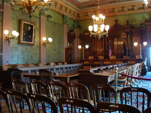 A historic courtroom with wooden furniture, ornate decor, and rows of chairs facing a large judge's bench.