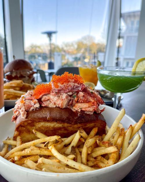A bowl of fries topped with a lobster roll, garnished with orange roe, with drinks and burgers in the background.
