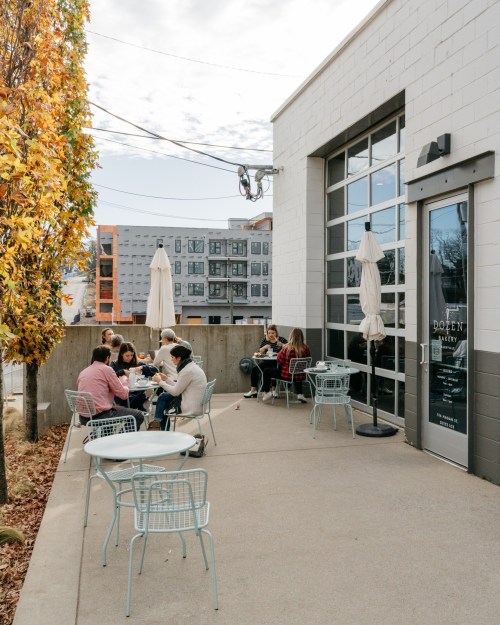 A cozy outdoor café scene with people dining at tables, surrounded by trees and modern buildings in the background.
