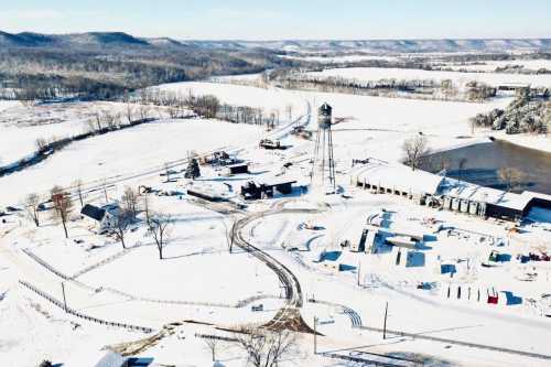 Aerial view of a snowy rural landscape with a water tower, buildings, and winding roads surrounded by hills.