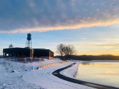 A snowy landscape at sunset featuring a building, a water tower, and a calm lake reflecting the sky.