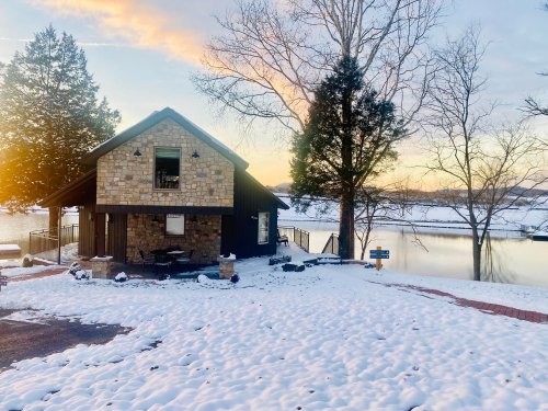 A cozy stone cabin by a snowy lake at sunset, surrounded by trees and a peaceful winter landscape.