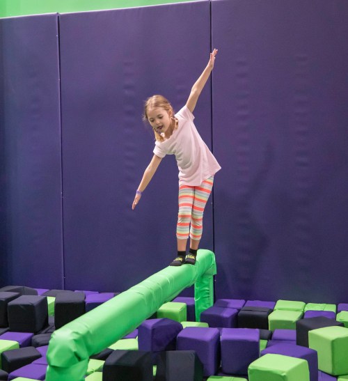 A young girl balances on a green beam above a pit filled with colorful foam blocks.