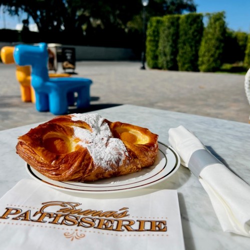 A pastry dusted with powdered sugar on a plate, with a napkin and a colorful chair in the background.