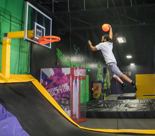 A person jumps on a trampoline, reaching for a basketball hoop while holding a ball in a vibrant indoor trampoline park.