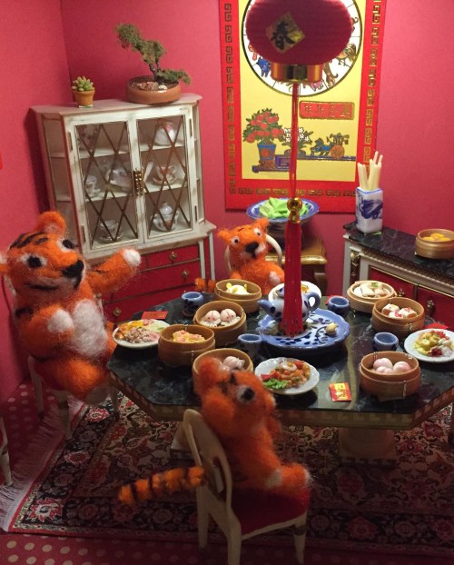 Three orange plush tigers sit around a dining table filled with food in a cozy, decorated room.