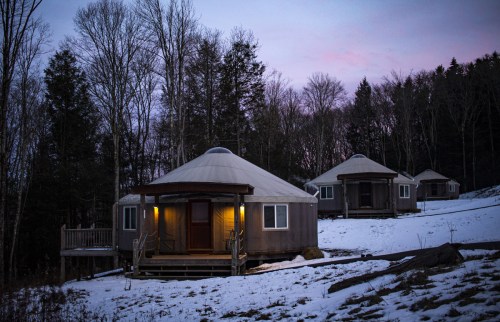 Three cozy yurts nestled in a snowy landscape, surrounded by trees at dusk, with warm lights glowing from the entrances.