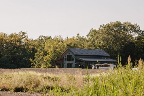 A modern barn-style building surrounded by trees and tall grass in a rural landscape.