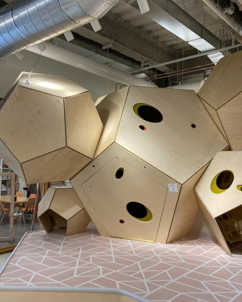 A large, geometric wooden play structure with yellow accents, set in a modern indoor space with a patterned floor.