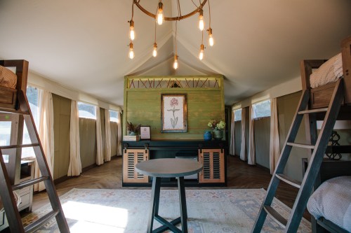 A cozy interior of a glamping tent featuring bunk beds, a round table, and decorative wall art under warm lighting.