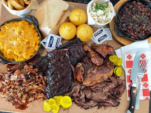 A large platter of barbecue meats, sides of mac and cheese, coleslaw, cornbread, pickles, and sauces.