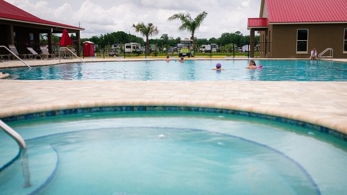 A swimming pool with people enjoying the water, surrounded by lounge chairs and palm trees under a cloudy sky.