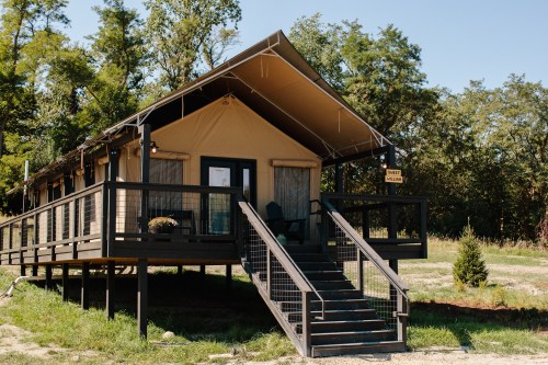 A cozy tent cabin on stilts, featuring a porch and stairs, surrounded by greenery and trees.
