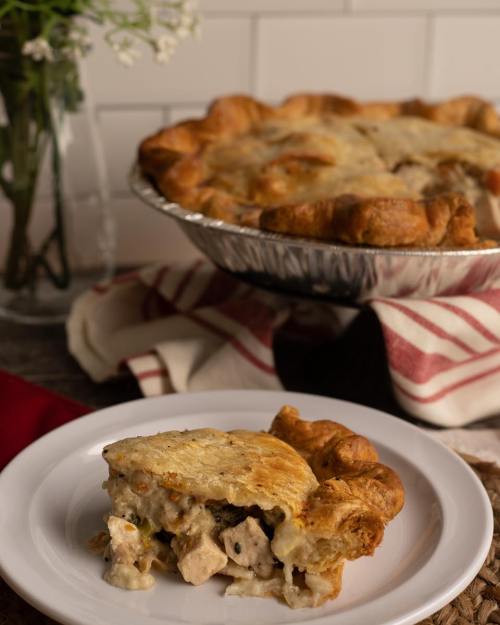 A slice of chicken pot pie on a plate, with the whole pie in the background and a vase of flowers nearby.