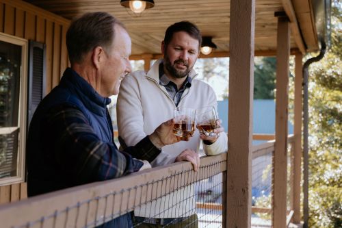 Two men on a balcony toast with glasses of whiskey, enjoying a moment together in a relaxed setting.
