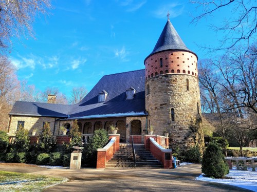 A stone building with a conical tower, surrounded by trees and a clear blue sky, with a dusting of snow on the ground.
