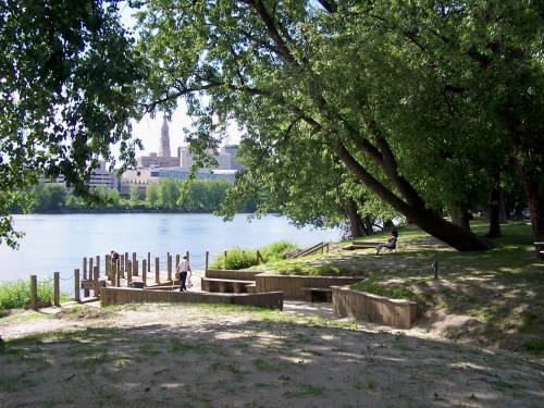A serene riverside scene with trees, a sandy area, and people enjoying the view of the water and city skyline.