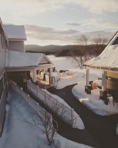 A snowy landscape with a path leading to a lake, surrounded by mountains and a cozy building in the foreground.