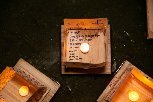 Wooden boxes with candles float on water, featuring handwritten wishes and messages on their sides.