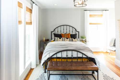 Cozy bedroom featuring a metal bed frame, patterned bedding, a wooden bench, and natural light from large windows.