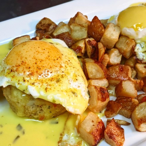 A plate of eggs Benedict with a poached egg on a muffin, served with crispy diced potatoes and hollandaise sauce.