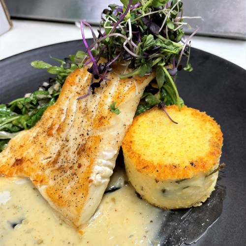 A plate featuring a piece of grilled fish, creamy sauce, a round potato cake, and a garnish of microgreens.