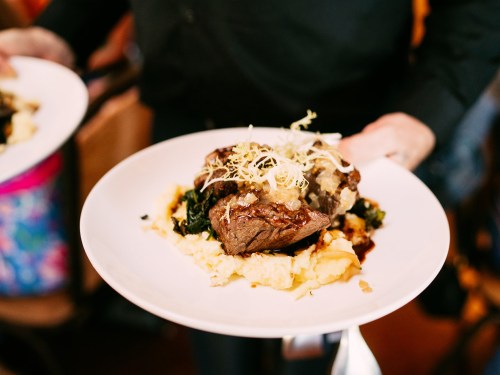 A server holds a plate with a serving of beef, mashed potatoes, and greens, garnished with herbs.