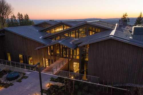 Modern wooden building with a unique roof design, surrounded by greenery, at sunset.