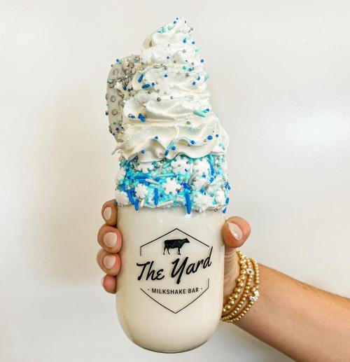 A hand holding a large milkshake topped with whipped cream, blue sprinkles, and a cookie, from The Yard Milkshake Bar.