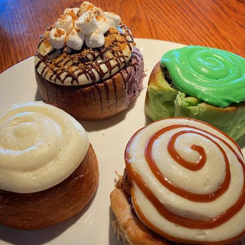 A plate of four colorful cinnamon rolls with various toppings, including chocolate, green icing, and marshmallows.