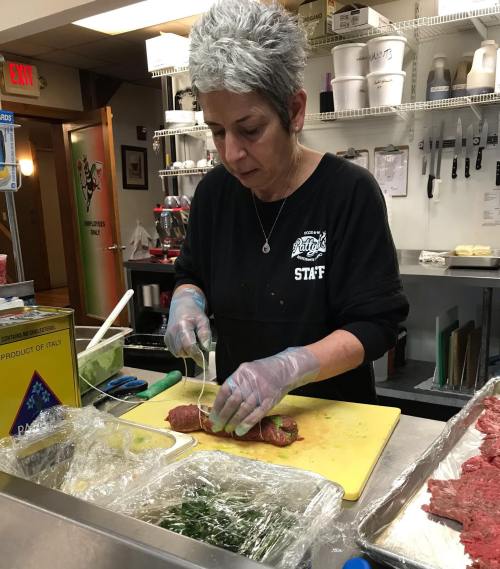 A woman with gray hair prepares food in a kitchen, slicing meat on a cutting board while wearing gloves.